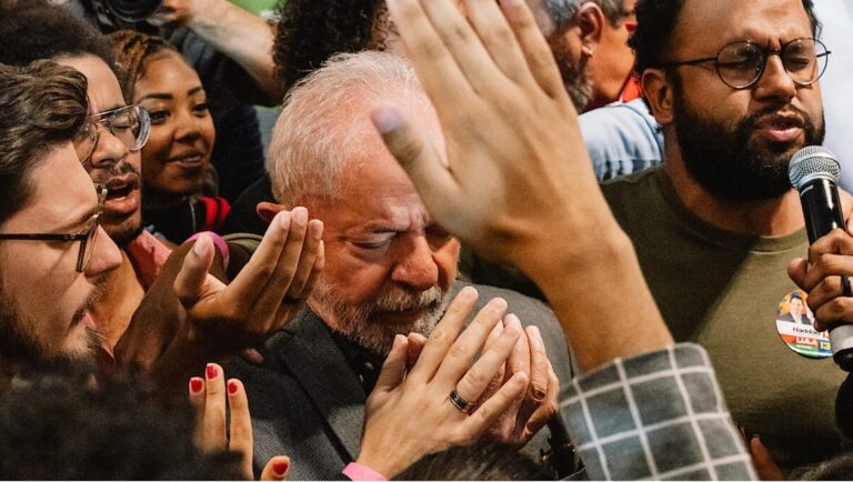 Luiz Inácio Lula da Silva surrounded by supporters. Lula, Brazilian politician, eyes closed, hands clasped. Crowd with raised hands.