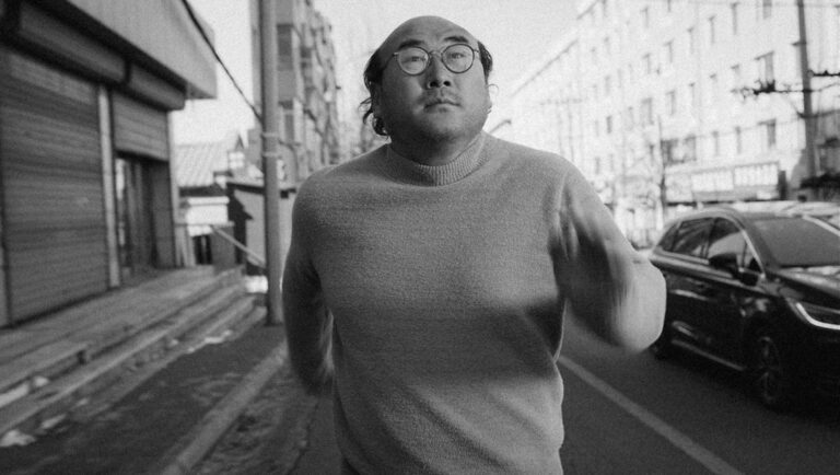Black and white film still. Man with glasses runs on street. Car and buildings in background. Bel-ami.