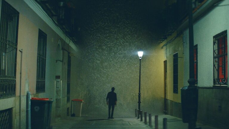 Night alleyway with a man walking toward a lit street lamp. Urban scene.