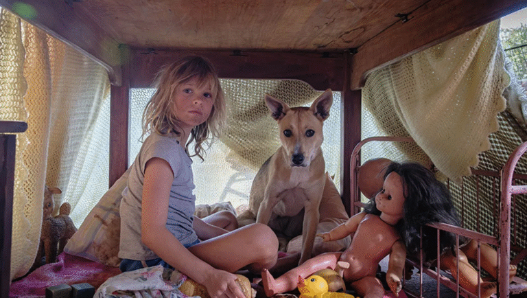 Girl, dog, and dolls inside a blanket fort. Childhood scene with toys and makeshift shelter.