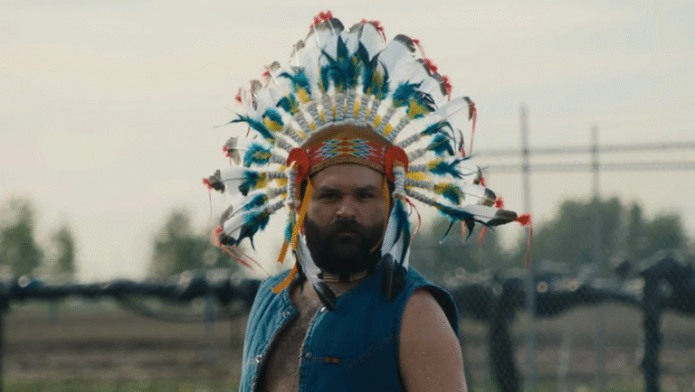 Man with beard wearing a colorful Native American headdress and blue vest.