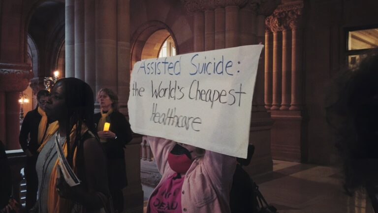 Protest against assisted suicide. Sign reads: Assisted Suicide: the World's Cheapest Healthcare. Activists present.