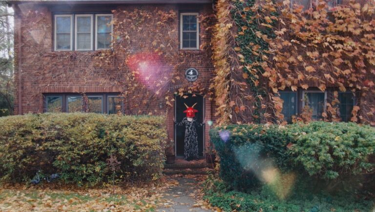 Brick house with vine foliage and person in doorway wearing a red devil mask and patterned robe.
