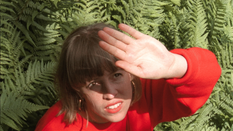 Film still from FIDMarseille 2025: A woman shields her eyes from the sun, ferns in background. Indie film.