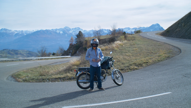 FIDMarseille 2025 film still: Man with helmet and motorcycle on winding mountain road. Snowy peaks in background.