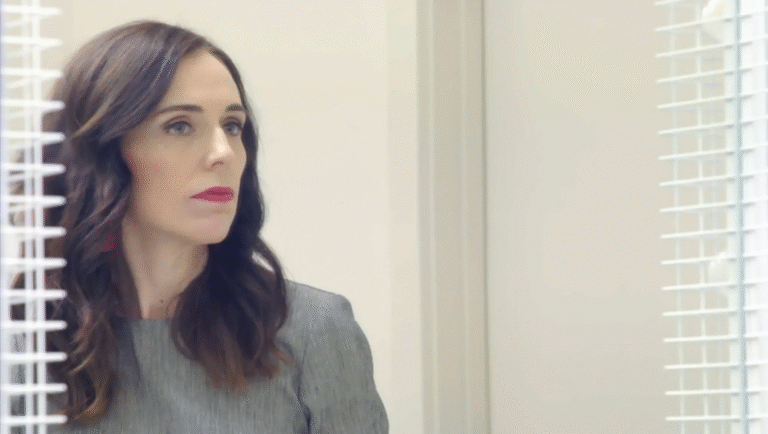 Jacinda Ardern, New Zealand's Prime Minister, looks to the side. She wears a gray suit and red lipstick.