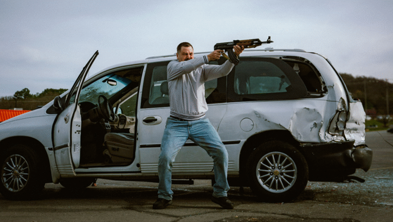 Man aims assault rifle by damaged minivan. Action scene, gun violence, crime.