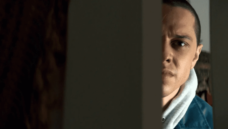 Man peeking through a doorway. Intense gaze, blue jacket, white hoodie. Suspenseful, dramatic lighting.