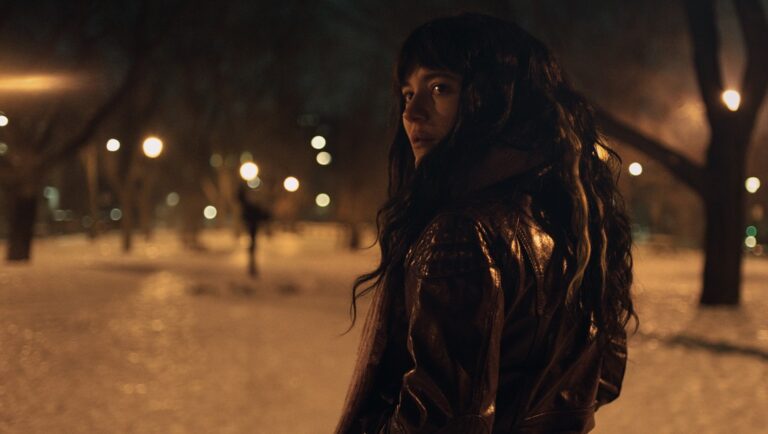 Woman in leather jacket looks back in snowy park at night. Moody, dark, cinematic.
