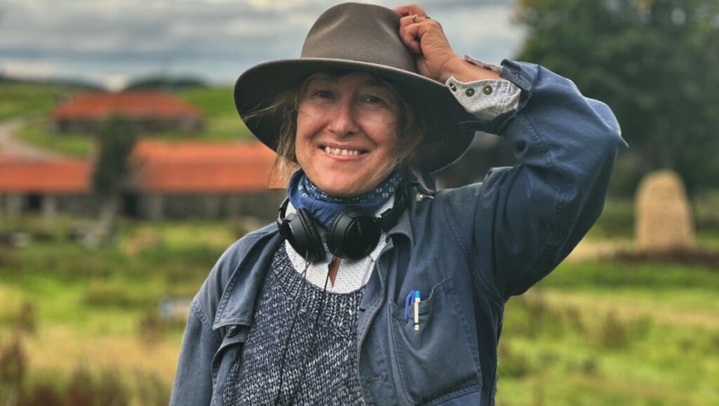Athina Rachel Tsangari headshot. Smiling woman with hat, headphones, and blue jacket in a rural landscape. Filmmaker portrait.