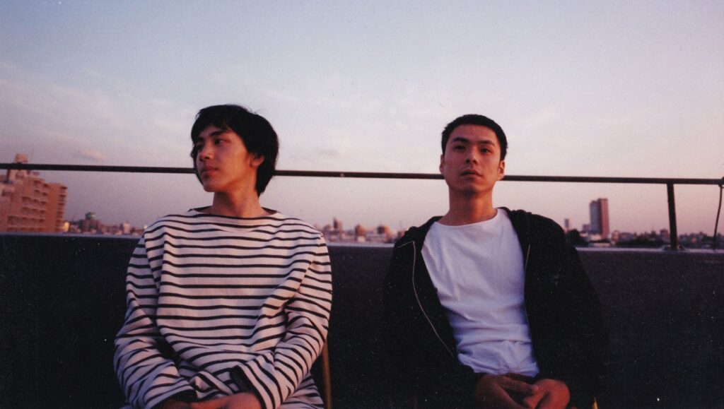 Two Asian men on a rooftop at sunset. Striped shirt, white tee, city skyline. Indie film aesthetic.