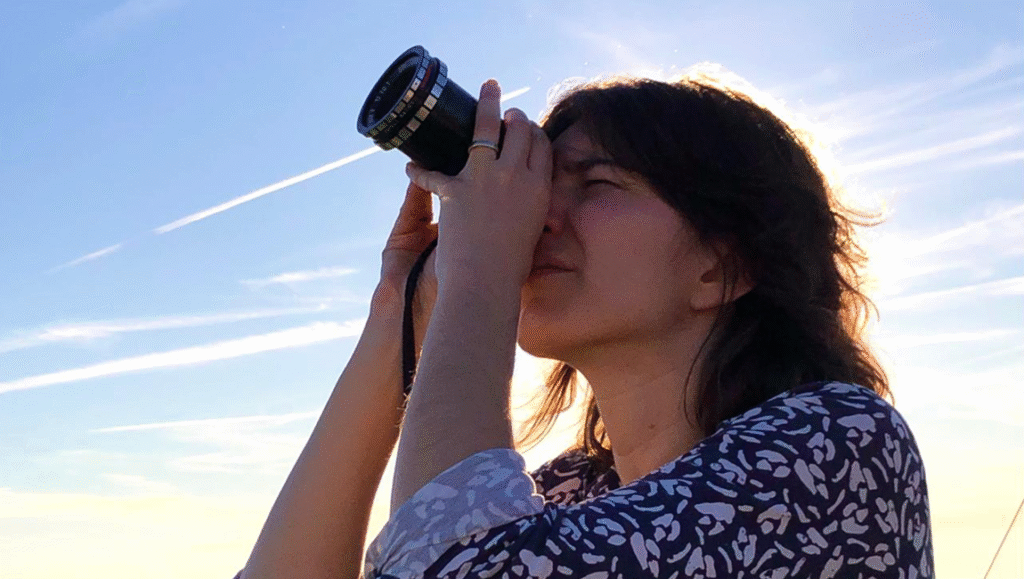 Woman looks through camera lens against a sunny sky. Photography, filmmaker, creative.