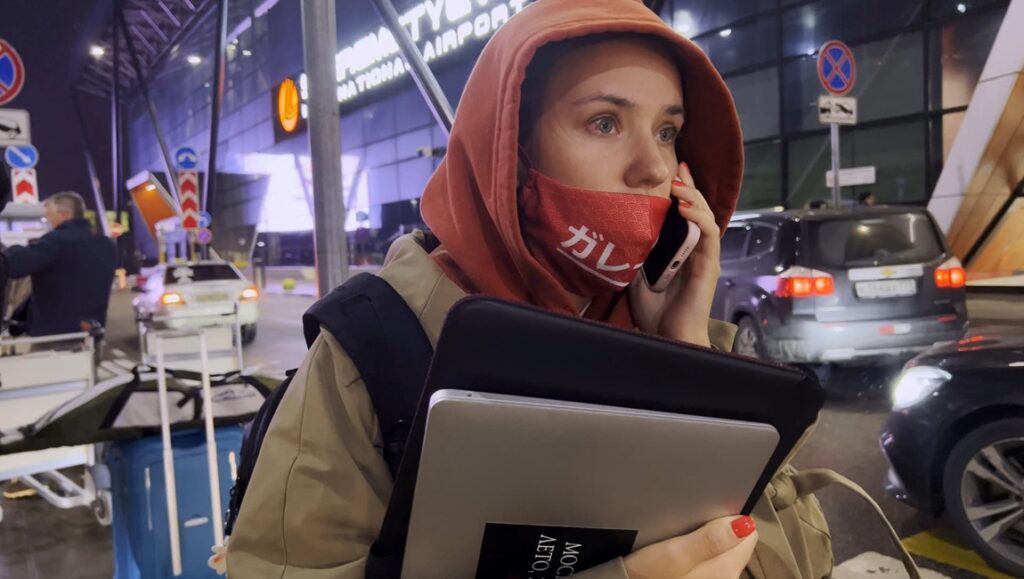 Woman at airport wearing mask, hoodie, and backpack. Talking on phone, holding laptop. Travel, airport scene.