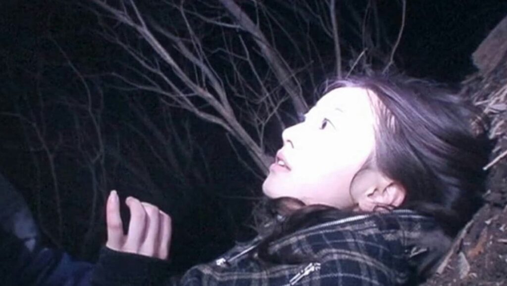 Noroi: The Curse movie scene. Young woman looks up at dark trees. Japanese horror film.
