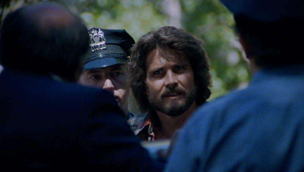 Night of the Juggler movie scene: a man with a beard and long hair is flanked by two police officers.