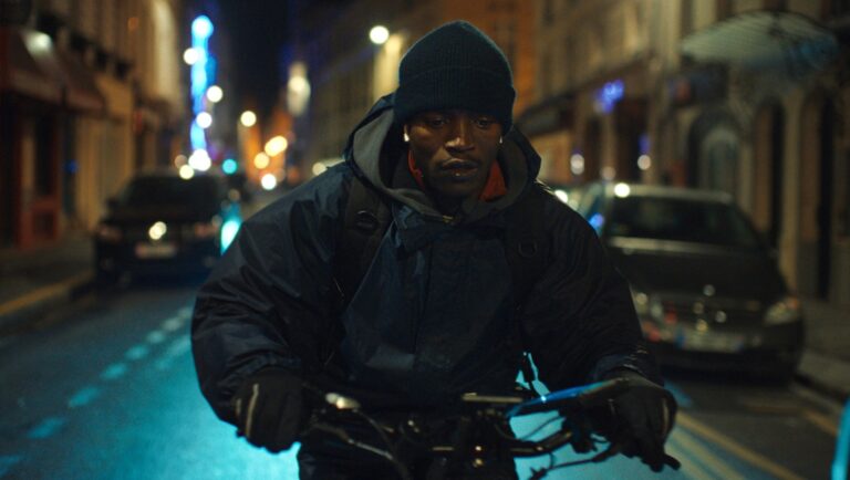 Man on scooter at night. Souleymane's Story. Urban commute. Dark street. Black beanie. Gloves. Electric scooter. City lights.