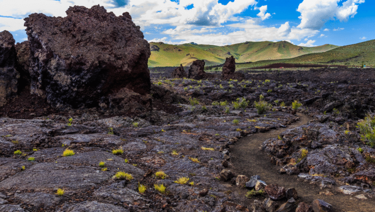 Idaho's Craters of the Moon: Black lava field, trail, mountains, and blue sky with clouds. Volcanic landscape.