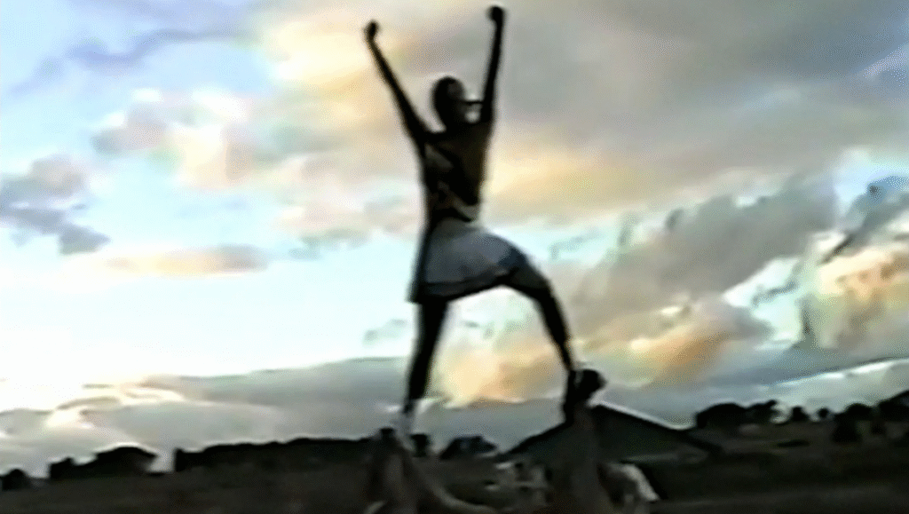 Some of You Fucked Up Eva film still. Cheerleader stands atop another person, arms raised, against a cloudy sky.