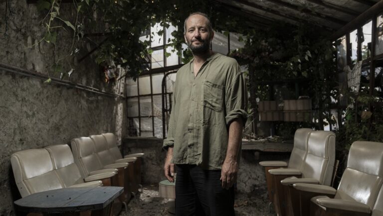 Man in overgrown greenhouse. Interior with chairs, plants, and natural light. Rustic, vintage aesthetic.